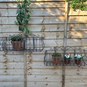 May include: Two wrought iron wall planters with a decorative scroll design. Each planter holds terracotta pots with plants. The planters are mounted on a wooden fence. Greenery is visible in the background.