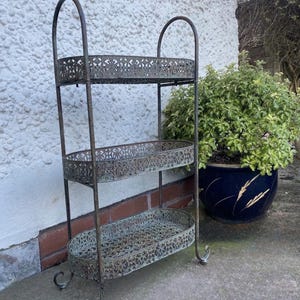 May include: A three-tiered metal plant stand with an ornate design. The stand is painted in a distressed green finish and has oval-shaped shelves. The stand is shown outdoors against a white brick wall.