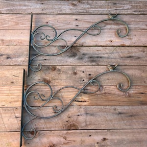 May include: Two dark metal wall brackets featuring ornate scrollwork designs and a small bird figure perched on the curved end of each arm. The brackets have a flat vertical mounting plate and a decorative, sweeping metal frame with curls.