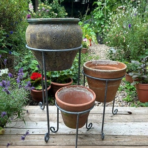 May include: Three terracotta plant pots on a metal stand. The largest pot is a mottled green and brown colour. The two smaller pots are a reddish brown colour. The stand is black metal with a decorative scroll design.