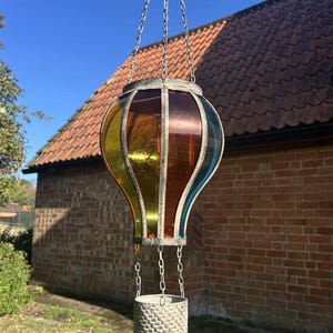 May include: A decorative hanging lantern in the shape of a hot air balloon. The lantern has panels of stained glass in yellow, orange, and blue, set within a metal frame. A woven basket is suspended below by chains. The background shows a brick building and blue sky.