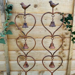 May include: Two rustic metal garden stakes with heart designs. Each stake has a small bird, a heart shape, and three bells. The stakes are set against a wooden fence background.