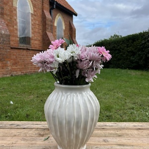 May include: A cream-coloured ceramic vase with vertical ribbed detailing holds a bouquet of pink and white flowers. The vase sits on a wooden surface, with a brick building and green lawn in the background.