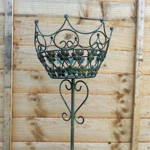 May include: An antique-style metal plant stand with a crown-shaped top and floral basket design. The stand is supported by a slender pole with heart-shaped accents. The finish is a weathered green, set against a wooden fence background. The stand's height is approximately 120 cm.