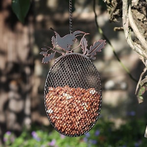 May include: A metal bird feeder shaped like a bird perched on a branch with leaves. The feeder is filled with peanuts and hangs from a tree branch.