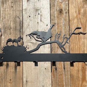 May include: Black metal wall-mounted coat rack featuring a silhouette of a bird carrying a twig in its beak and three baby birds in a nest. The rack has six hooks.