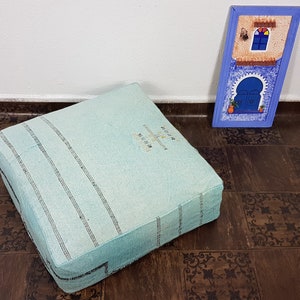 May include: A light blue square floor cushion with white stripes and a white embroidered design. The cushion is sitting on a brown floor with a decorative pattern.