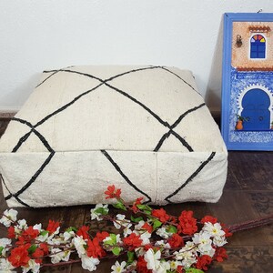 Puede incluir: Un coj&iacute;n de suelo blanco con un patr&oacute;n de diamante negro, sentado en un suelo de madera con una imagen enmarcada azul de una puerta marroqu&iacute; detr&aacute;s.