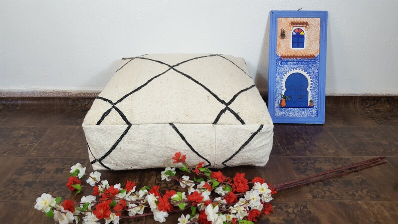 Puede incluir: Un coj&iacute;n de suelo blanco con un patr&oacute;n de diamante negro, sentado en un suelo de madera con una imagen enmarcada azul de una puerta marroqu&iacute; detr&aacute;s.