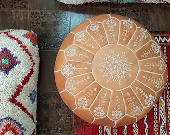 May include: A tan leather pouf with white embroidery, featuring a starburst design in the centre. The pouf is sitting on a red and white patterned rug.