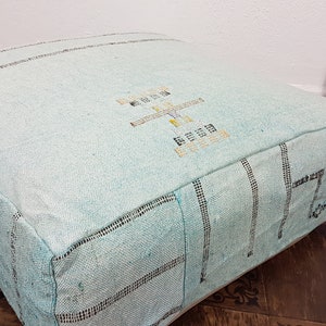 May include: A light blue square pouf with a woven pattern and embroidered design. The pouf is made of a soft, textured fabric.