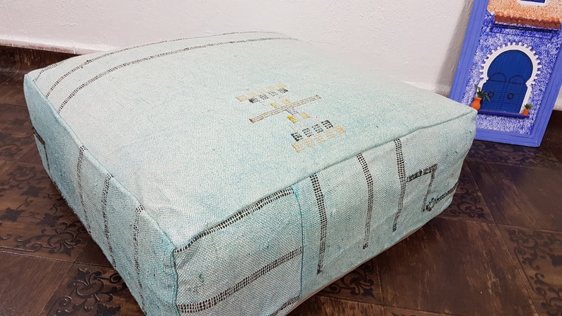 May include: A light blue square pouf with a woven pattern and embroidered design. The pouf is made of a soft, textured fabric.