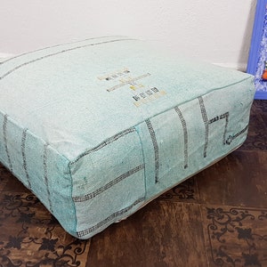 May include: A light blue square pouf with a white and black geometric pattern. The pouf is made of woven fabric and has a soft, comfortable texture.