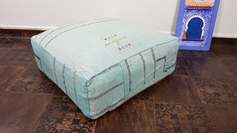 May include: A light blue square pouf with a white and black geometric pattern. The pouf is made of woven fabric and has a soft, comfortable texture.