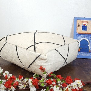 Puede incluir: Un coj&iacute;n de suelo blanco y negro con estampado geom&eacute;trico y contorno negro. El coj&iacute;n est&aacute; sobre un suelo de madera con un peque&ntilde;o cuadro azul enmarcado que representa un edificio con una puerta y una ventana azules en el fondo.