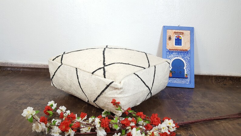 Puede incluir: Un coj&iacute;n de suelo blanco y negro con estampado geom&eacute;trico y contorno negro. El coj&iacute;n est&aacute; sobre un suelo de madera con un peque&ntilde;o cuadro azul enmarcado que representa un edificio con una puerta y una ventana azules en el fondo.