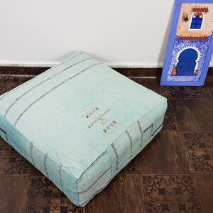 May include: A light blue square pouf with a woven pattern and embroidered design. The pouf is sitting on a brown tiled floor.