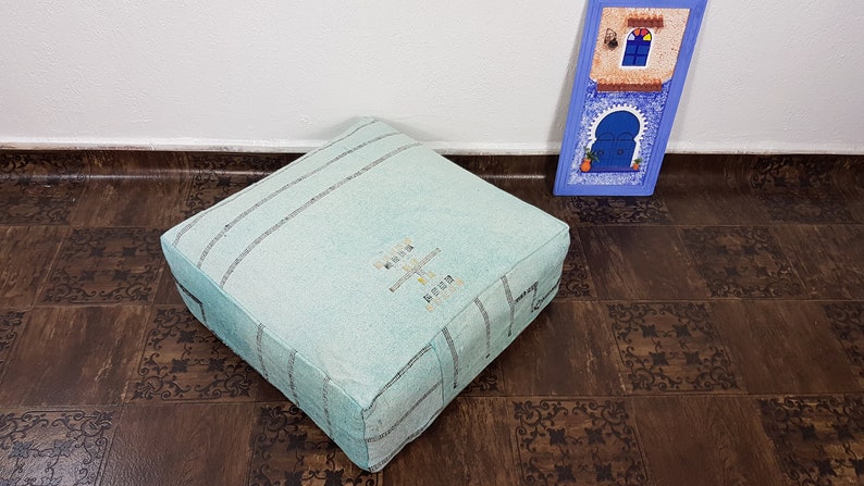 May include: A light blue square pouf with a woven pattern and embroidered design. The pouf is sitting on a brown tiled floor.