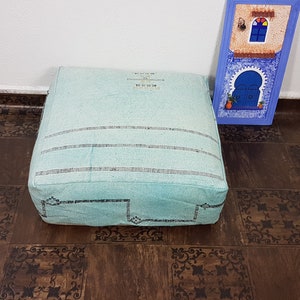 May include: A light blue square floor cushion with black embroidered details. The cushion is sitting on a brown tiled floor.