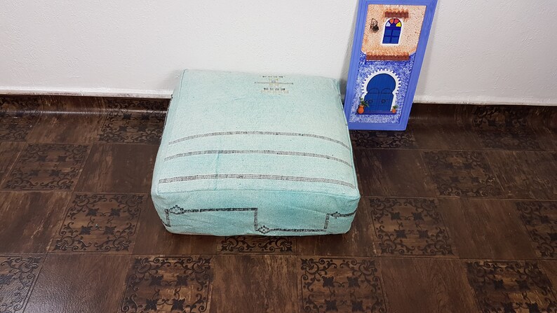 May include: A light blue square floor cushion with black embroidered details. The cushion is sitting on a brown tiled floor.