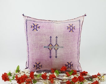 May include: A soft, pink throw pillow with a geometric pattern in shades of purple, blue, and yellow. The pillow is made of a woven fabric and has a fringed edge.