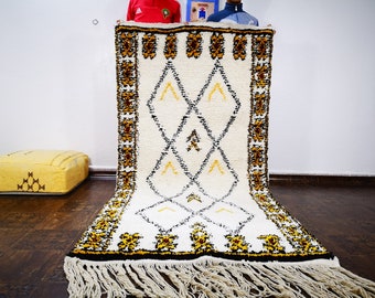 May include: A white Moroccan rug with a geometric pattern in black, brown, and yellow. The rug has a fringe border and is being held up by two people.
