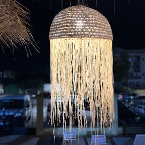 May include: A woven, light brown, dome-shaped lampshade with a fringe of long, thin strands hanging down. The lampshade is lit from within, casting a warm glow.