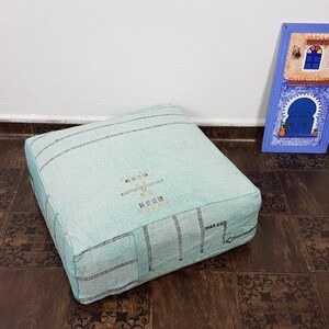 May include: A light blue square floor cushion with a white embroidered design. The cushion is sitting on a brown patterned floor.