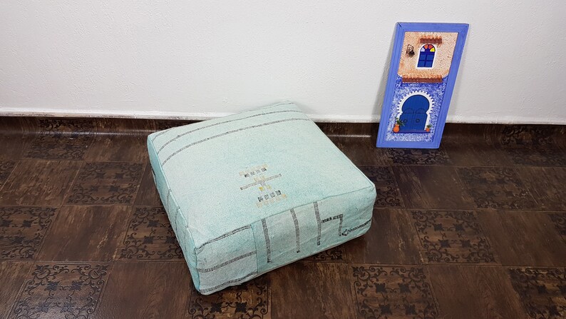 May include: A light blue square floor cushion with a white embroidered design. The cushion is sitting on a brown patterned floor.