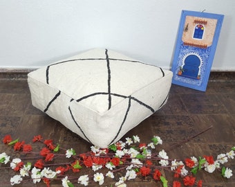 Op de afbeelding: Een wit vloer kussen met zwarte geometrische ontwerpen. Het kussen ligt op een bruine houten vloer met een klein blauw fotolijstje op de achtergrond.