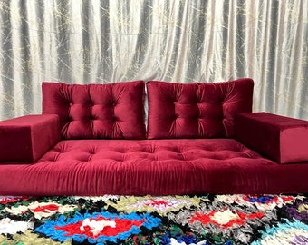 May include: A deep red velvet sofa with tufted cushions and armrests. The sofa is sitting on a colourful rug with a variety of patterns.