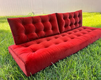 May include: A red velvet futon sofa with tufted cushions and a thick mattress. The sofa is sitting on a green grassy lawn.