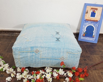 Pode incluir: Um pufe quadrado azul claro com desenhos geométricos bordados. O pufe é feito de um tecido texturizado e tem costuras decorativas. Uma obra de arte emoldurada com uma porta e uma janela azuis está ao fundo.