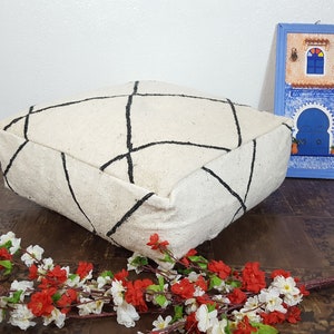 Puede incluir: Un coj&iacute;n de suelo blanco con un patr&oacute;n geom&eacute;trico negro. El coj&iacute;n est&aacute; sentado en un suelo de madera con un arreglo floral en primer plano.