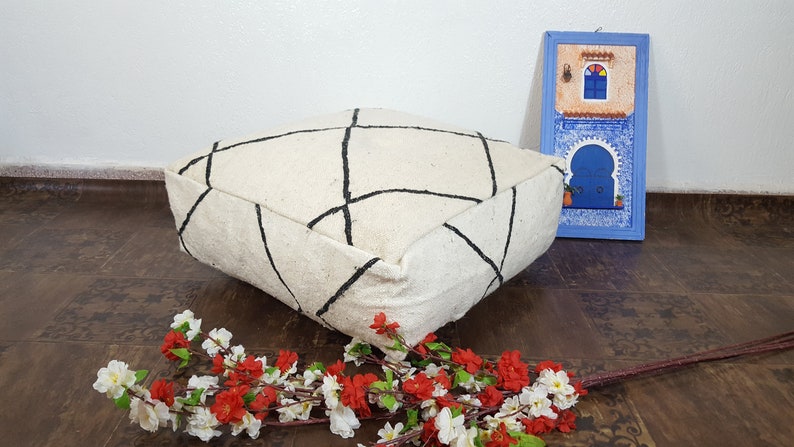 Puede incluir: Un coj&iacute;n de suelo blanco con un patr&oacute;n geom&eacute;trico negro. El coj&iacute;n est&aacute; sentado en un suelo de madera con un arreglo floral en primer plano.