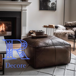 May include: A brown leather ottoman with a tufted design sits in a living room with a fireplace and a chair. The ottoman is in front of a coffee table with a white top. The word "Decore" is printed in blue on the floor in front of the ottoman.