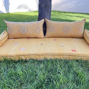 May include: A yellow Moroccan pouf with embroidered details and two matching pillows. The pouf is sitting on a green lawn.