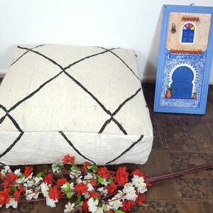 Puede incluir: Un coj&iacute;n de suelo blanco con un patr&oacute;n de diamante negro, una obra de arte enmarcada azul y blanca y un ramo de flores blancas y rojas sobre un suelo de patr&oacute;n marr&oacute;n.