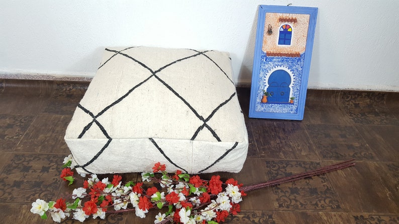 Puede incluir: Un coj&iacute;n de suelo blanco con un patr&oacute;n de diamante negro, una obra de arte enmarcada azul y blanca y un ramo de flores blancas y rojas sobre un suelo de patr&oacute;n marr&oacute;n.