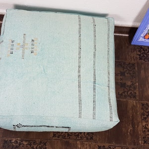 May include: A light blue square floor cushion with a woven pattern and a decorative design in the center. The cushion is sitting on a brown patterned floor.