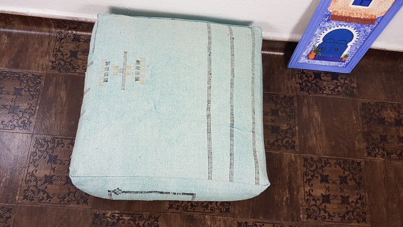 May include: A light blue square floor cushion with a woven pattern and a decorative design in the center. The cushion is sitting on a brown patterned floor.
