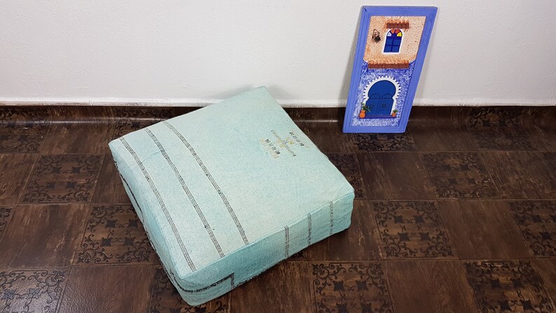 May include: A light blue square floor cushion with white stripes and a white embroidered design. The cushion is sitting on a brown floor with a decorative pattern.