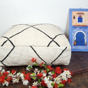 Puede incluir: Un coj&iacute;n de suelo blanco con un patr&oacute;n de diamantes negros, sentado en un suelo de madera con una imagen enmarcada azul de una puerta marroqu&iacute; y un ramo de flores blancas y rojas.