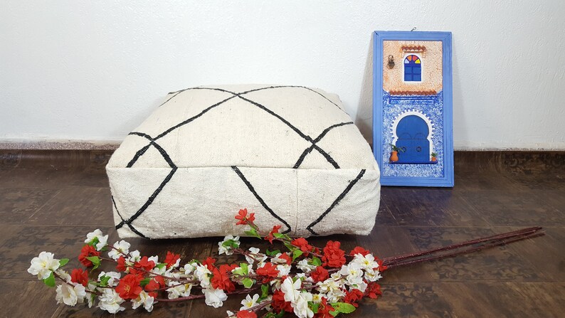 Puede incluir: Un coj&iacute;n de suelo blanco con un patr&oacute;n de diamantes negros, sentado en un suelo de madera con una imagen enmarcada azul de una puerta marroqu&iacute; y un ramo de flores blancas y rojas.