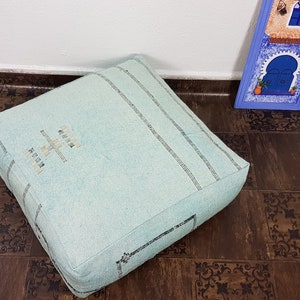 May include: A light blue square floor cushion with black embroidered stripes and the words "Made in Morocco" in white. The cushion is sitting on a brown patterned floor.