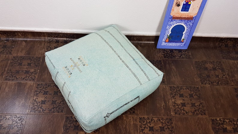 May include: A light blue square floor cushion with black embroidered stripes and the words "Made in Morocco" in white. The cushion is sitting on a brown patterned floor.