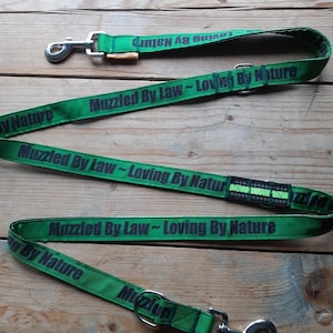 May include: Green dog lead with black text that reads "Muzzled By Law - Loving By Nature". The lead has a silver clip on each end.