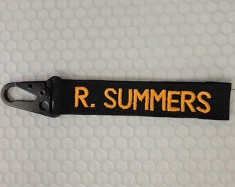 May include: A black fabric keychain strap featuring the embroidered text R. SUMMERS in yellow thread. The strap is attached to a matte black metal carabiner clip with a spring-loaded gate mechanism on the left side.