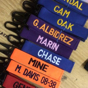 May include: A stack of colorful fabric keychains with embroidered names, including "Bug's Ba", "Kendal", "Cam Oak", "G. Albidrez", "Marin", "Chase", "Mine", "M. Davis 08-38", "Genevish", and "J. Hue".