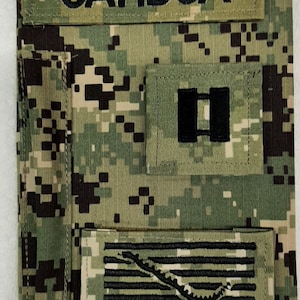 May include: A military uniform patch with the name "GAMBOA" in black letters. The patch features a camouflage pattern and includes a rank insignia and a "Don't Tread on Me" emblem.